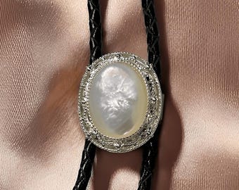 May include: An oval bolo tie pendant with a pearlescent center and a silver-colored, ornate border. The pendant hangs from two black, braided leather cords. The background is a soft, peach-colored fabric.