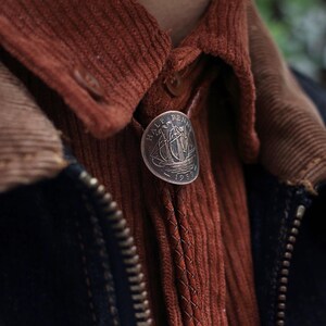 May include: A vintage-style bolo tie featuring a 1959 Half Penny coin with a sailing ship design. The coin is set against a rust-colored corduroy shirt and a dark blue jacket, creating a classic, rustic aesthetic.