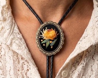 May include: A black bolo tie with a gold-toned oval pendant featuring a yellow flower design. The pendant is framed by a decorative border. The tie has a black braided cord.