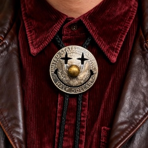 May include: A silver-toned bolo tie featuring a smiling face design with a gold-colored ball in the center. The tie is made from a United States half dollar coin. The bolo tie is worn over a burgundy corduroy shirt and a brown leather jacket.