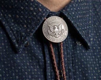 May include: A silver Half Dollar coin bolo tie with a brown leather cord. The coin features an eagle and the words "United States of America" and "Half Dollar". The tie is worn with a dark blue button-down shirt.