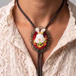 May include: A bolo tie featuring a dark brown braided leather cord and an oval-shaped antique bronze pendant. The pendant contains a red velvet background with an embroidered white rabbit surrounded by small orange and green floral accents.