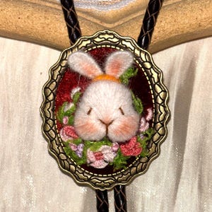 May include: A bolo tie featuring a central oval pendant with an antique bronze scalloped frame. The pendant contains a needle-felted white rabbit head surrounded by embroidered pink and green flowers on a deep red background.