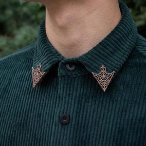 May include: A dark green corduroy shirt with decorative bronze collar pins. The pins feature an intricate, symmetrical pattern. The shirt has a button-down front and a classic collar. The background is blurred, suggesting an outdoor setting.