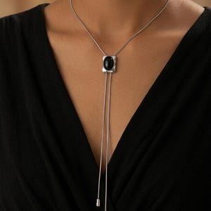 以下が含まれることがあります： 黒い楕円形の石のペンダントが付いた、シルバーカラーのネックレス。ペンダントは長方形で、中央に黒い石があります。2本のシルバーチェーンがペンダントから垂れ下がり、それぞれ小さな丸みを帯びたシルバーのチャームで終わっています。ネックレスは黒いトップスの上に着用されています。