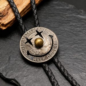 May include: A silver half-dollar coin bolo tie with a black braided leather cord. The coin features a smiley face design with a gold-colored center. The coin is engraved with the words "UNITED STATES OF AMERICA" and "HALF DOLLAR".