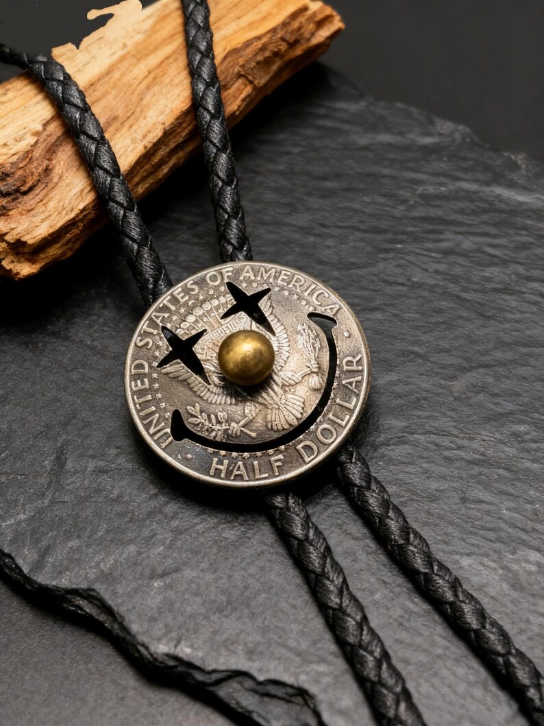 May include: A silver half-dollar coin bolo tie with a black braided leather cord. The coin features a smiley face design with a gold-colored center. The coin is engraved with the words "UNITED STATES OF AMERICA" and "HALF DOLLAR".