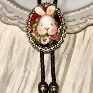 May include: A bolo tie featuring a central oval bronze-toned frame containing a needle-felted white rabbit head surrounded by embroidered flowers on a red background. The tie has a braided black cord with bronze-toned metal tips engraved with True Love.