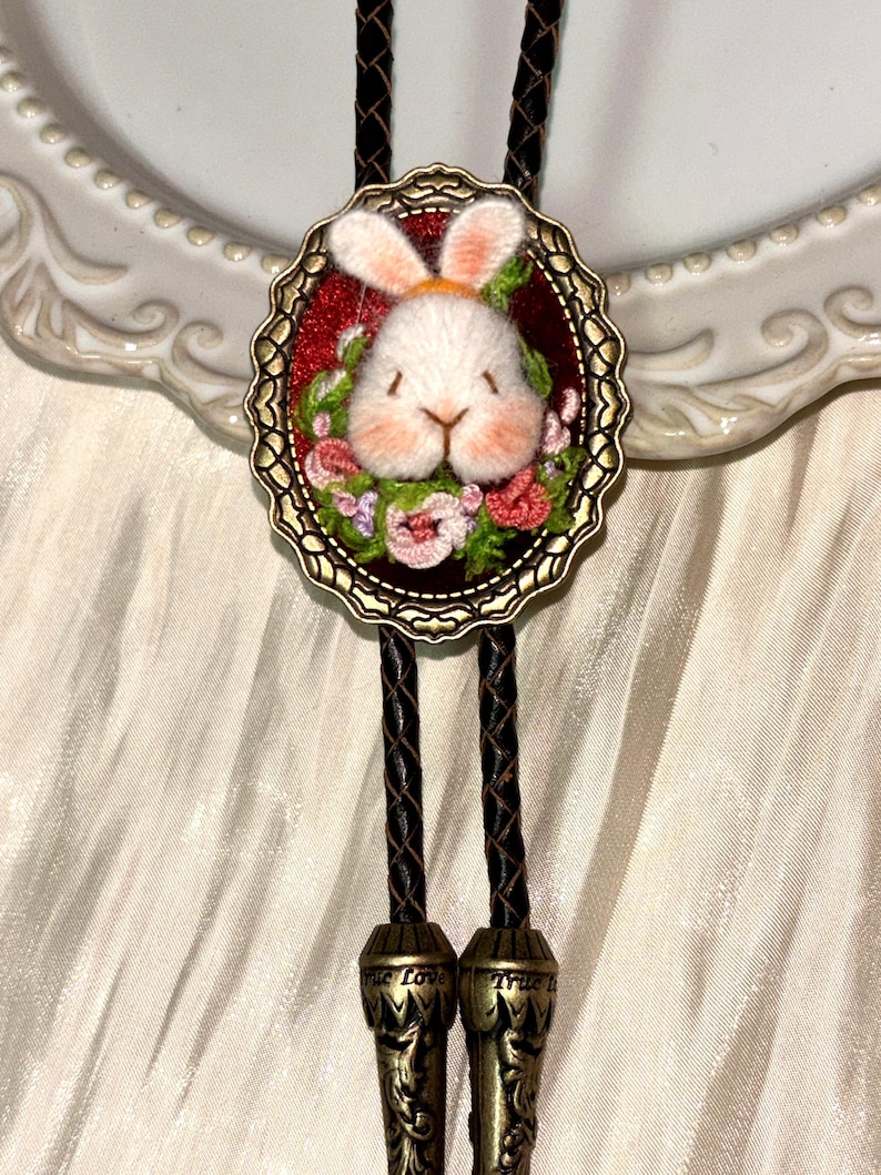 May include: A bolo tie featuring a central oval bronze-toned frame containing a needle-felted white rabbit head surrounded by embroidered flowers on a red background. The tie has a braided black cord with bronze-toned metal tips engraved with True Love.