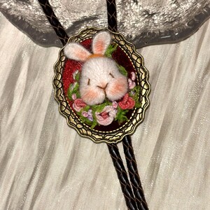 May include: A bolo tie featuring a dark brown braided leather cord and an oval antique bronze frame. The center contains a needle-felted white rabbit head with pink accents, surrounded by small embroidered pink and green floral details.