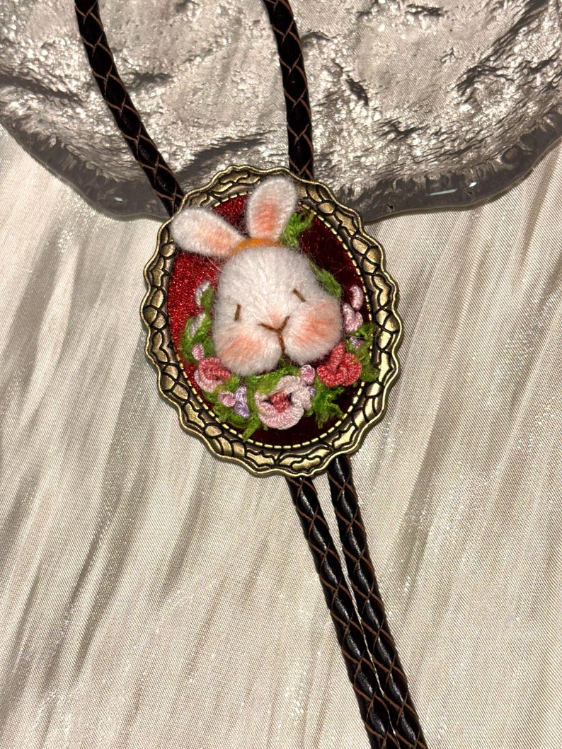 May include: A bolo tie featuring a dark brown braided leather cord and an oval antique bronze frame. The center contains a needle-felted white rabbit head with pink accents, surrounded by small embroidered pink and green floral details.