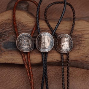 Könnte beinhalten: Drei Vintage-Bolo-Krawatten mit Münzen. Die Bolo-Krawatten haben braune, schwarze und braune Lederkordeln. Die runden Münzen zeigen detaillierte Designs. Eine Münze zeigt ein Schiff, und die anderen beiden zeigen ein Profil.