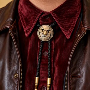 May include: A bronze bolo tie with a circular pendant featuring a decorative design. The pendant hangs from a black braided cord with gold-colored tips. The tie is worn over a burgundy corduroy shirt and a brown leather jacket.