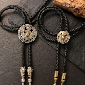 May include: Two bolo ties with black braided cords and silver-toned and brass-toned metal accents. One bolo tie features a silver-toned coin with a smiley face design. The other has a similar design.
