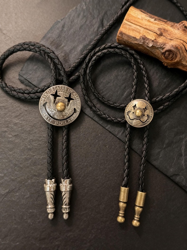 May include: Two bolo ties with black braided cords and silver-toned and brass-toned metal accents. One bolo tie features a silver-toned coin with a smiley face design. The other has a similar design.