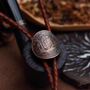 May include: A copper-toned bolo tie featuring a 1959 Half Penny coin with a sailing ship design. The coin is attached to a brown braided leather cord. The tie rests on a dark, textured surface, with a pipe in the background.