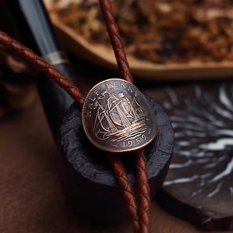 May include: A copper-toned bolo tie featuring a 1959 Half Penny coin with a sailing ship design. The coin is attached to a brown braided leather cord. The tie rests on a dark, textured surface, with a pipe in the background.