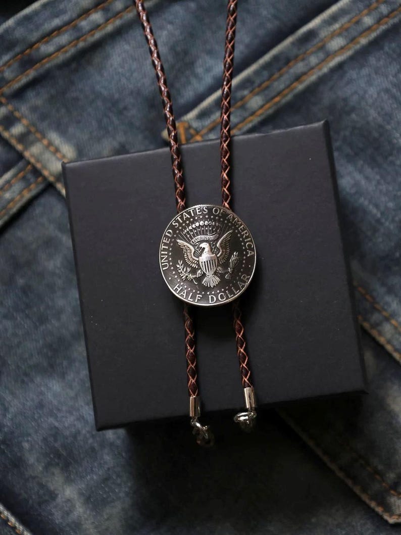 K&ouml;nnte beinhalten: Silberfarbene Bolo-Krawatte mit einer US-amerikanischen Half-Dollar-M&uuml;nze mit Adler-Emblem. Die M&uuml;nze ist an einer braunen geflochtenen Lederschnur befestigt. Die Krawatte liegt auf einer schwarzen Schachtel vor Jeansstoff.