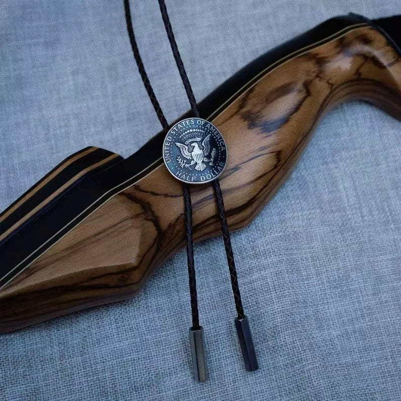 K&ouml;nnte beinhalten: Eine Bolo-Krawatte mit einer silbernen Half-Dollar-M&uuml;nze, einer dunkelbraunen Lederschnur und Metallspitzen. Die M&uuml;nze zeigt das Siegel der Vereinigten Staaten von Amerika. Der Hintergrund ist aus Holz.