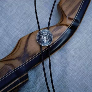 K&ouml;nnte beinhalten: Ein Bolo-Krawatte mit einer Silber-Halbdollar-M&uuml;nze, die das Siegel der Vereinigten Staaten von Amerika zeigt. Die M&uuml;nze ist an einem schwarzen geflochtenen Kordel befestigt, vor einem Holz Hintergrund mit schwarzen Akzenten.