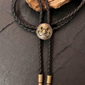 May include: A black braided leather bolo tie with a circular antique brass-colored pendant. The pendant has a raised central design and the words "REPUBLIQUE FRANCAISE" around the edge. The tie has brass-colored tips.