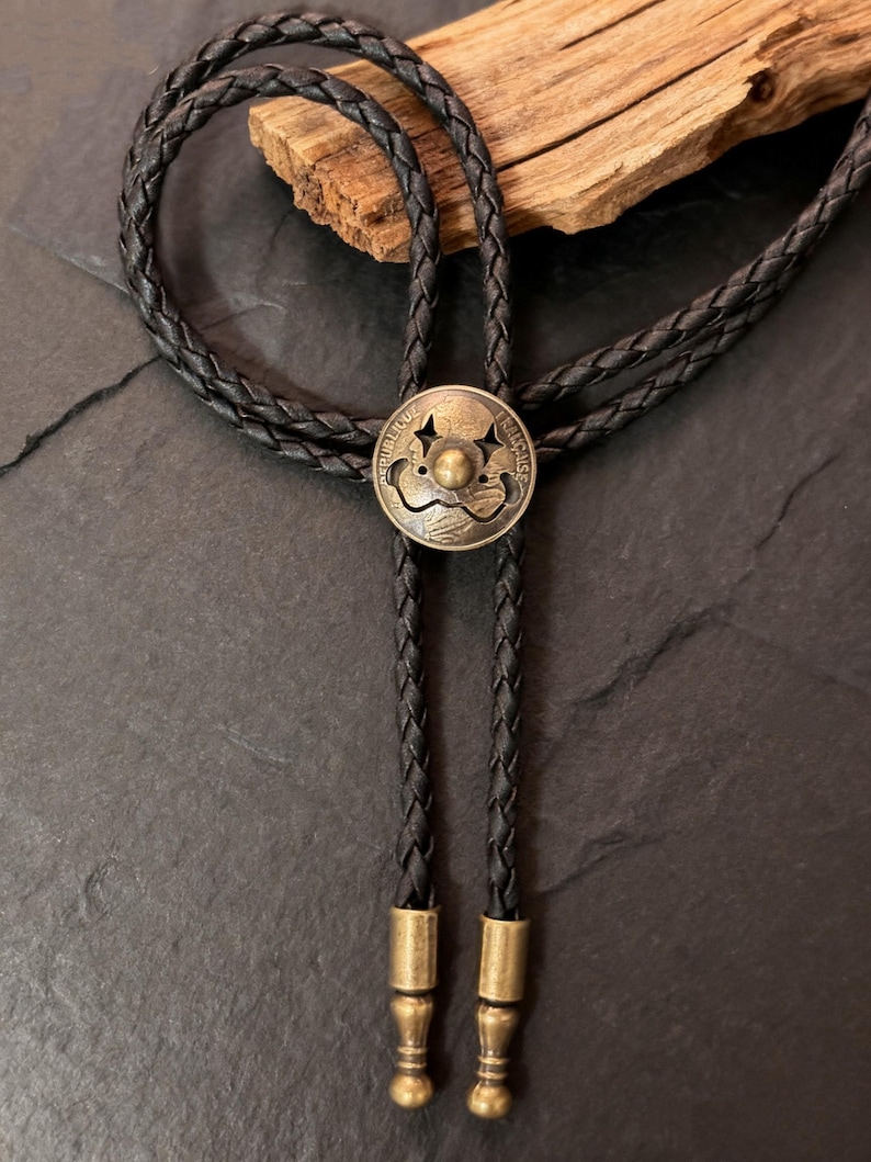 May include: A black braided leather bolo tie with a circular antique brass-colored pendant. The pendant has a raised central design and the words "REPUBLIQUE FRANCAISE" around the edge. The tie has brass-colored tips.