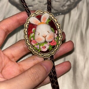 May include: A bolo tie featuring an oval antique brass frame with a scalloped edge. Inside the frame is a needle-felted white rabbit head with pink cheeks, surrounded by small embroidered green leaves and pink flowers on a dark red background.