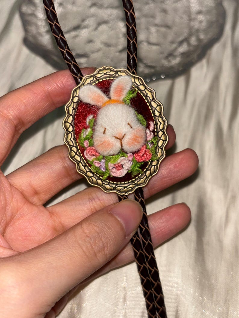 May include: A bolo tie featuring an oval antique brass frame with a scalloped edge. Inside the frame is a needle-felted white rabbit head with pink cheeks, surrounded by small embroidered green leaves and pink flowers on a dark red background.
