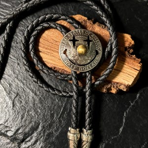 May include: A black braided leather bolo tie with a silver-toned half-dollar coin pendant. The coin features the words "UNITED STATES OF AMERICA" and "HALF DOLLAR". The tie has decorative silver-toned tips.