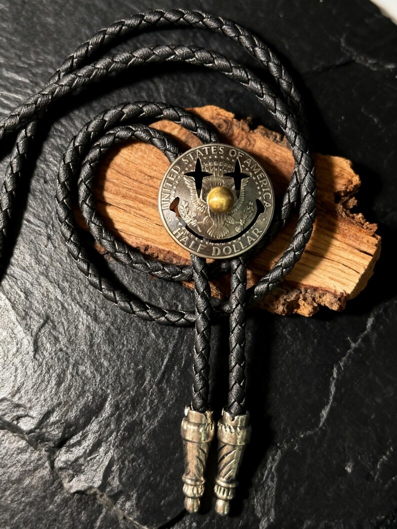 May include: A black braided leather bolo tie with a silver-toned half-dollar coin pendant. The coin features the words "UNITED STATES OF AMERICA" and "HALF DOLLAR". The tie has decorative silver-toned tips.