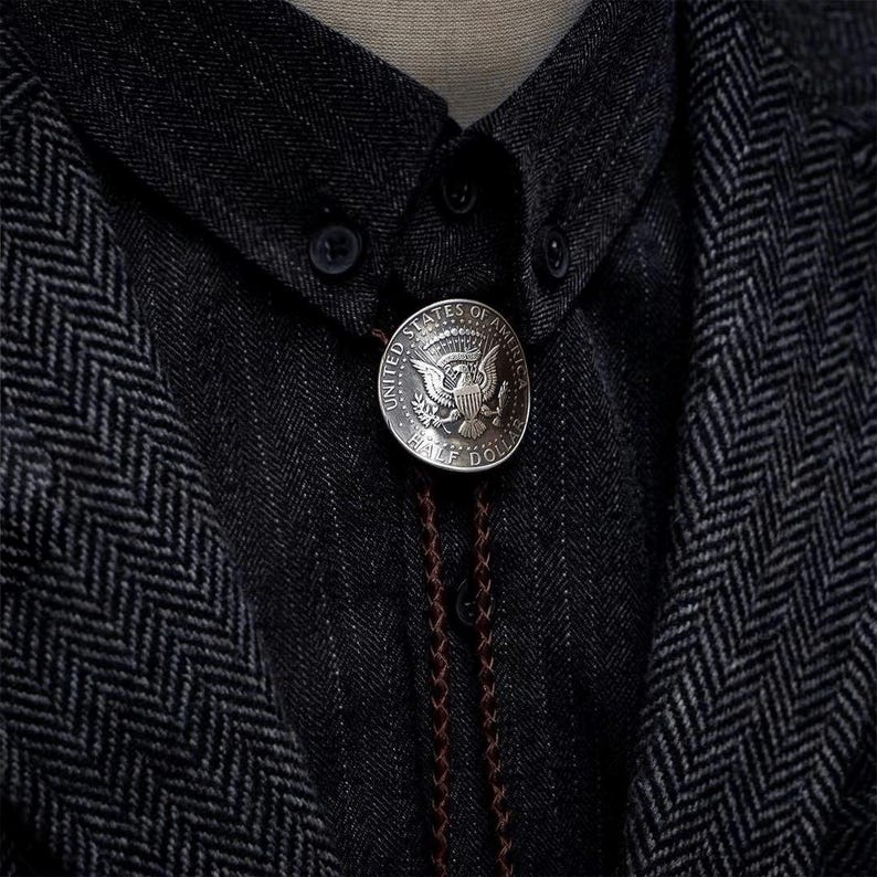 K&ouml;nnte beinhalten: Eine silberne Bolo-Krawatte mit einem Design einer Half-Dollar-M&uuml;nze der Vereinigten Staaten von Amerika. Die M&uuml;nze ist an einer braunen geflochtenen Kordel befestigt, vor einem dunkelgrauen Hemd und einer Jacke mit Fischgr&auml;tenmuster. Ein klassisches Accessoire.