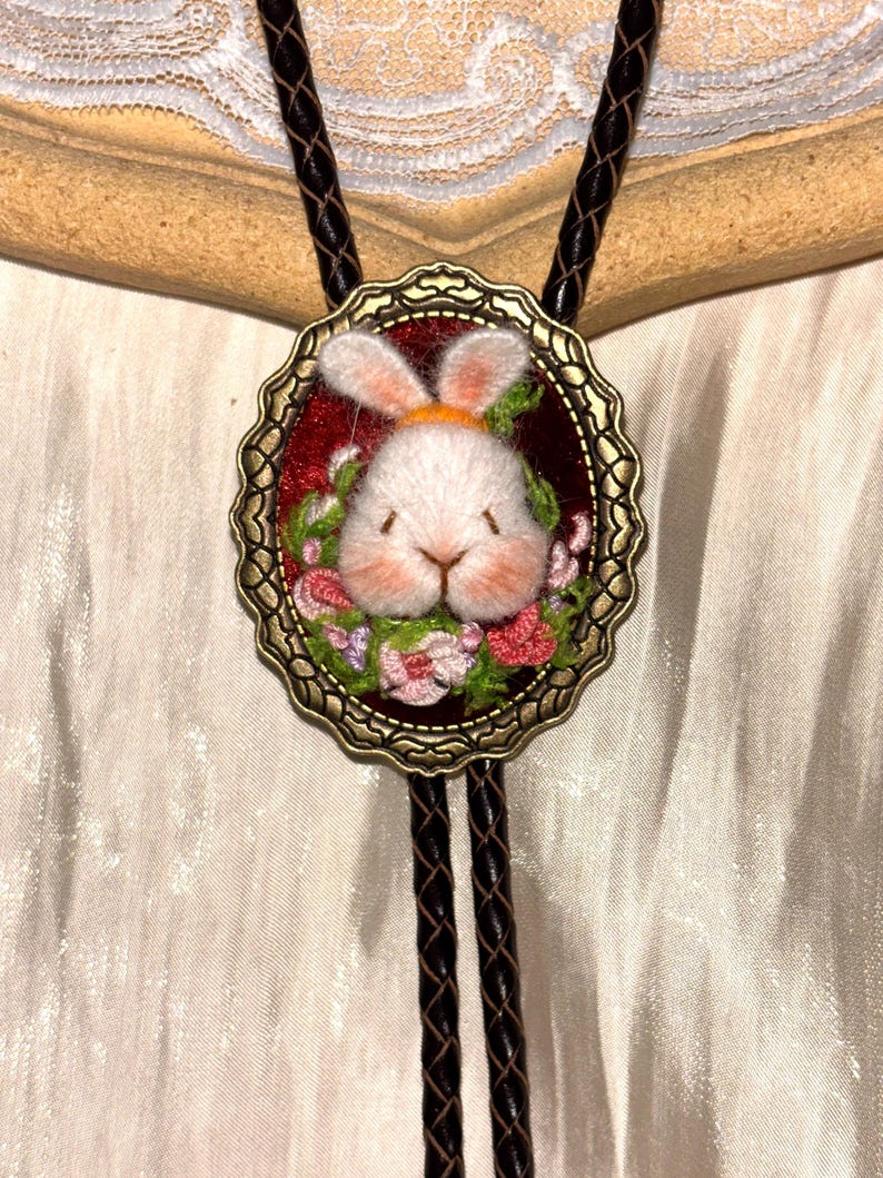 May include: A bolo tie featuring a central oval pendant with an antique bronze scalloped frame. The pendant contains a needle-felted white rabbit head surrounded by embroidered pink and green flowers on a deep red background.