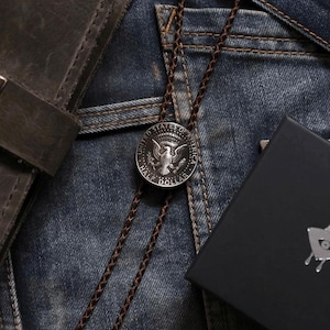 K&ouml;nnte beinhalten: Eine Bolo-Krawatte mit einer silbernen Half-Dollar-M&uuml;nze und einem braunen geflochtenen Kordel. Die M&uuml;nze zeigt ein Adlerdesign. Die Krawatte liegt auf einer Jeans. Eine schwarze Schachtel und eine Ledergeldb&ouml;rse sind ebenfalls im Bild.