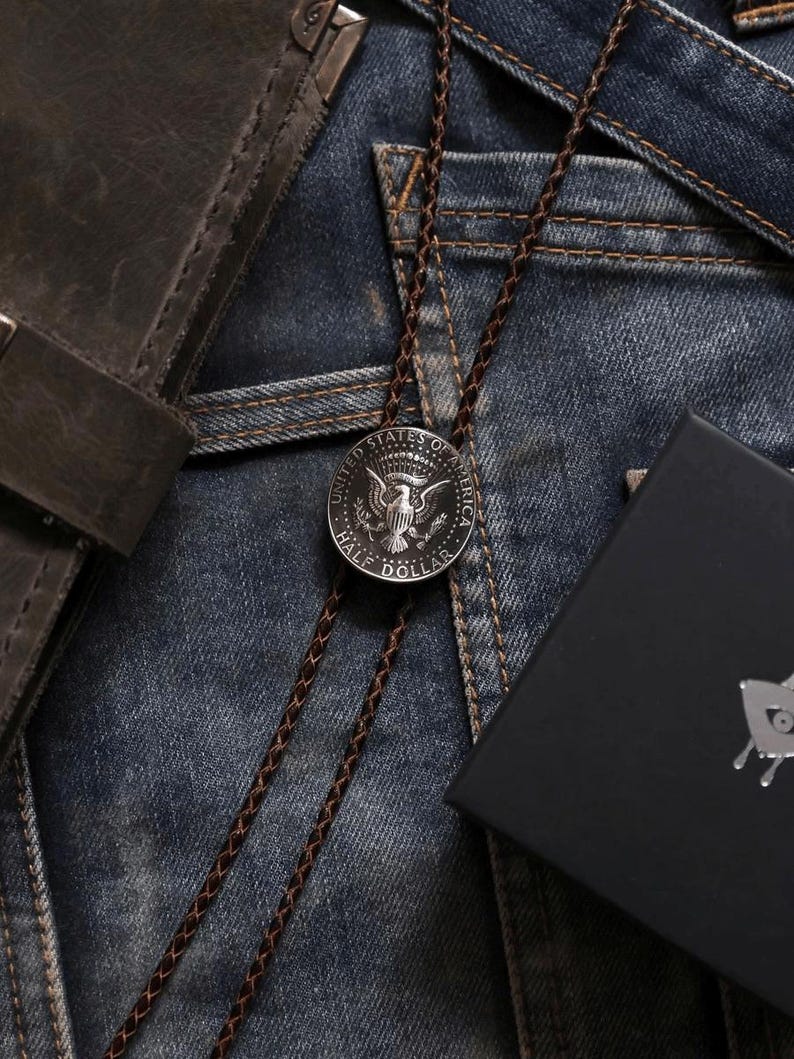 K&ouml;nnte beinhalten: Eine Bolo-Krawatte mit einer silbernen Half-Dollar-M&uuml;nze und einem braunen geflochtenen Kordel. Die M&uuml;nze zeigt ein Adlerdesign. Die Krawatte liegt auf einer Jeans. Eine schwarze Schachtel und eine Ledergeldb&ouml;rse sind ebenfalls im Bild.