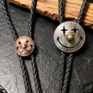 May include: Two bolo ties with black braided cords. One tie features a copper-colored circular pendant with a clown face design. The other has a silver half-dollar coin pendant with a smiley face and the words "UNITED STATES OF AMERICA HALF DOLLAR".
