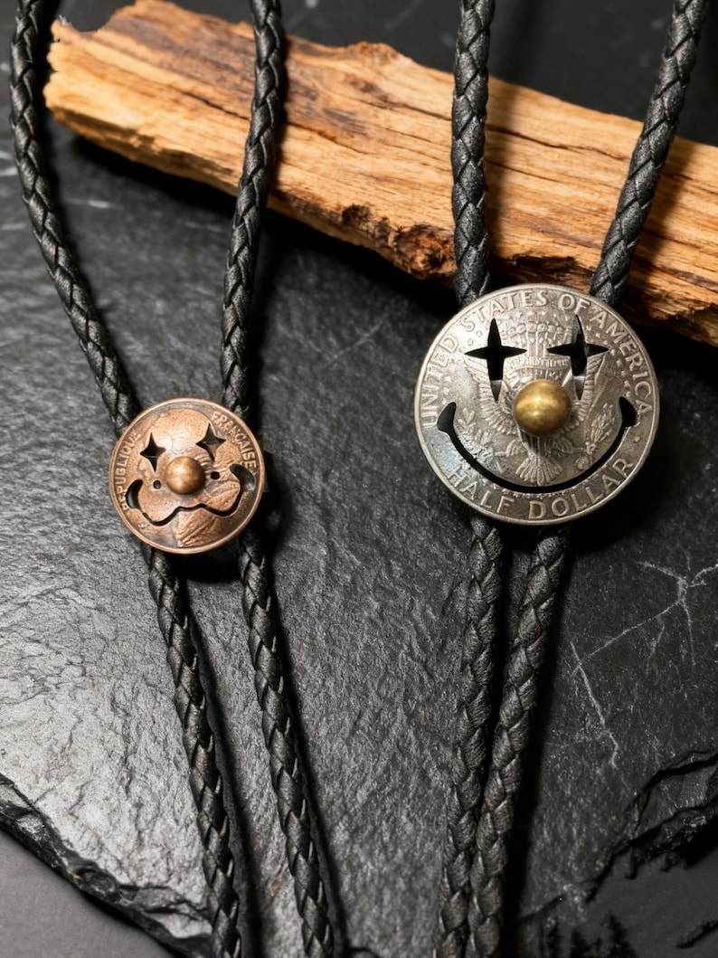 May include: Two bolo ties with black braided cords. One tie features a copper-colored circular pendant with a clown face design. The other has a silver half-dollar coin pendant with a smiley face and the words "UNITED STATES OF AMERICA HALF DOLLAR".