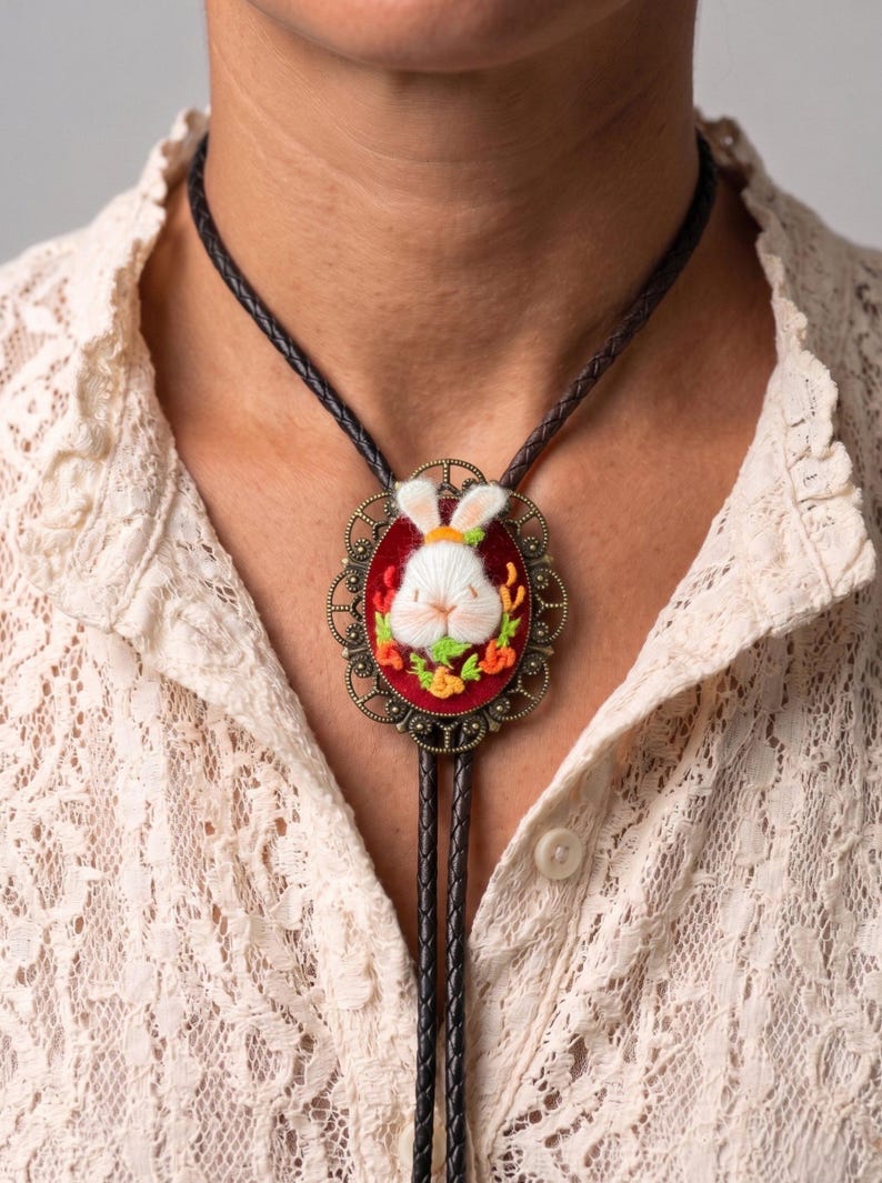 May include: A bolo tie featuring a dark brown braided leather cord and an oval-shaped antique bronze pendant. The pendant contains a red velvet background with an embroidered white rabbit surrounded by small orange and green floral accents.