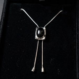 Flat lay of silver bolo tie necklace with black stone, highlighting clean lines, minimalist western design, and fine chain detail, ideal for modern women’s fashion and layering jewelry looks.