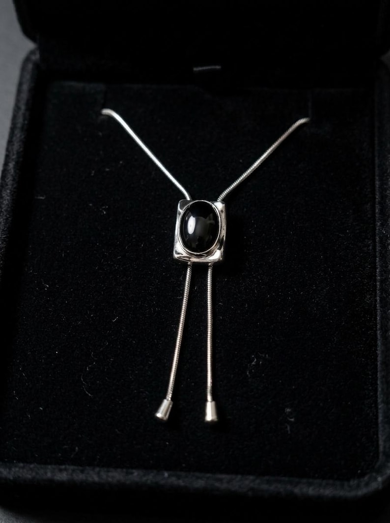 Flat lay of silver bolo tie necklace with black stone, highlighting clean lines, minimalist western design, and fine chain detail, ideal for modern women’s fashion and layering jewelry looks.