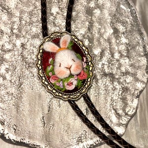 May include: A bolo tie featuring an oval antique brass frame containing a needle-felted white rabbit head with pink cheeks, surrounded by embroidered pink and green flowers on a dark red background, attached to a braided dark brown cord.