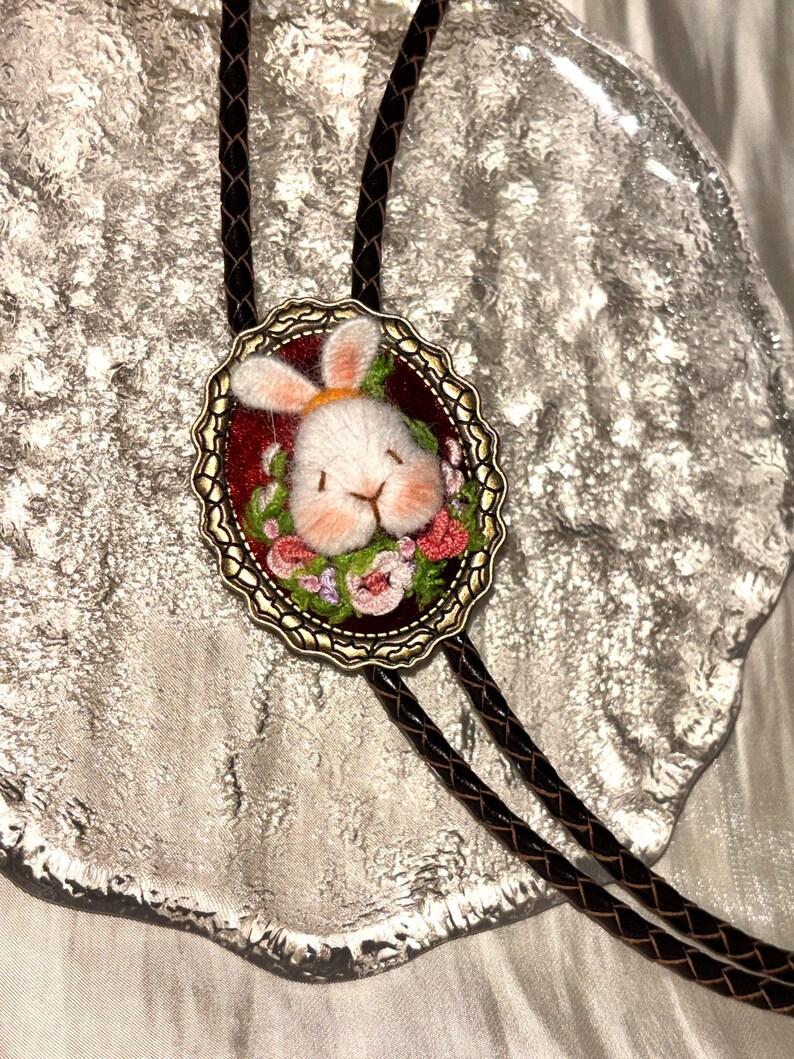 May include: A bolo tie featuring an oval antique brass frame containing a needle-felted white rabbit head with pink cheeks, surrounded by embroidered pink and green flowers on a dark red background, attached to a braided dark brown cord.