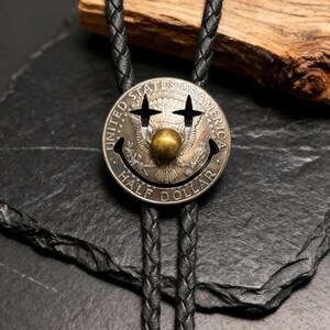 May include: A silver Half Dollar coin bolo tie with a smiley face design. The coin features cut-out eyes and a brass ball nose. The black braided leather cord is visible against a dark background.