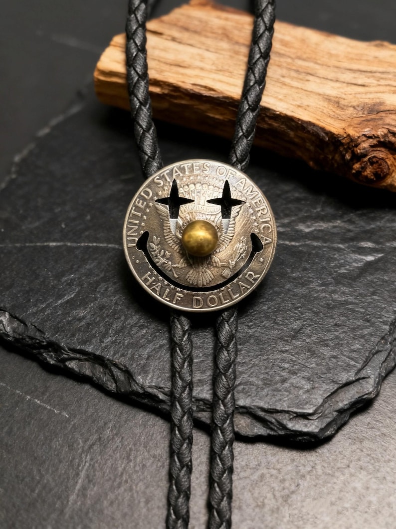 May include: A silver Half Dollar coin bolo tie with a smiley face design. The coin features cut-out eyes and a brass ball nose. The black braided leather cord is visible against a dark background.