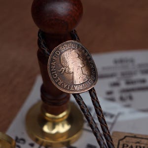 May include: A vintage-style bolo tie featuring a copper-toned coin with an embossed profile and inscription. The coin is attached to a braided dark brown leather cord. The tie is set against a blurred background.