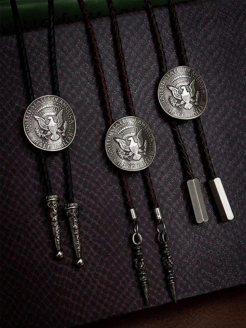 K&ouml;nnte beinhalten: Nahaufnahme von drei Bolo-Krawatten mit silbernen Half-Dollar-M&uuml;nzanh&auml;ngern. Die M&uuml;nzen tragen die Aufschrift "UNITED STATES OF AMERICA" und "HALF DOLLAR". Die Krawatten haben schwarze und braune geflochtene Kordeln mit Metallspitzen.