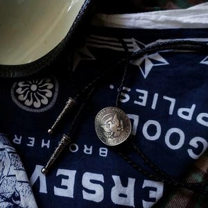 K&ouml;nnte beinhalten: Eine Bolo-Krawatte mit einer silbernen Half-Dollar-M&uuml;nze und einer schwarzen Lederschnur. Die M&uuml;nze zeigt einen Adler und die Worte "United States of America". Die Krawatte liegt auf einem marineblauen Bandana mit wei&szlig;em Text und Sternmustern.