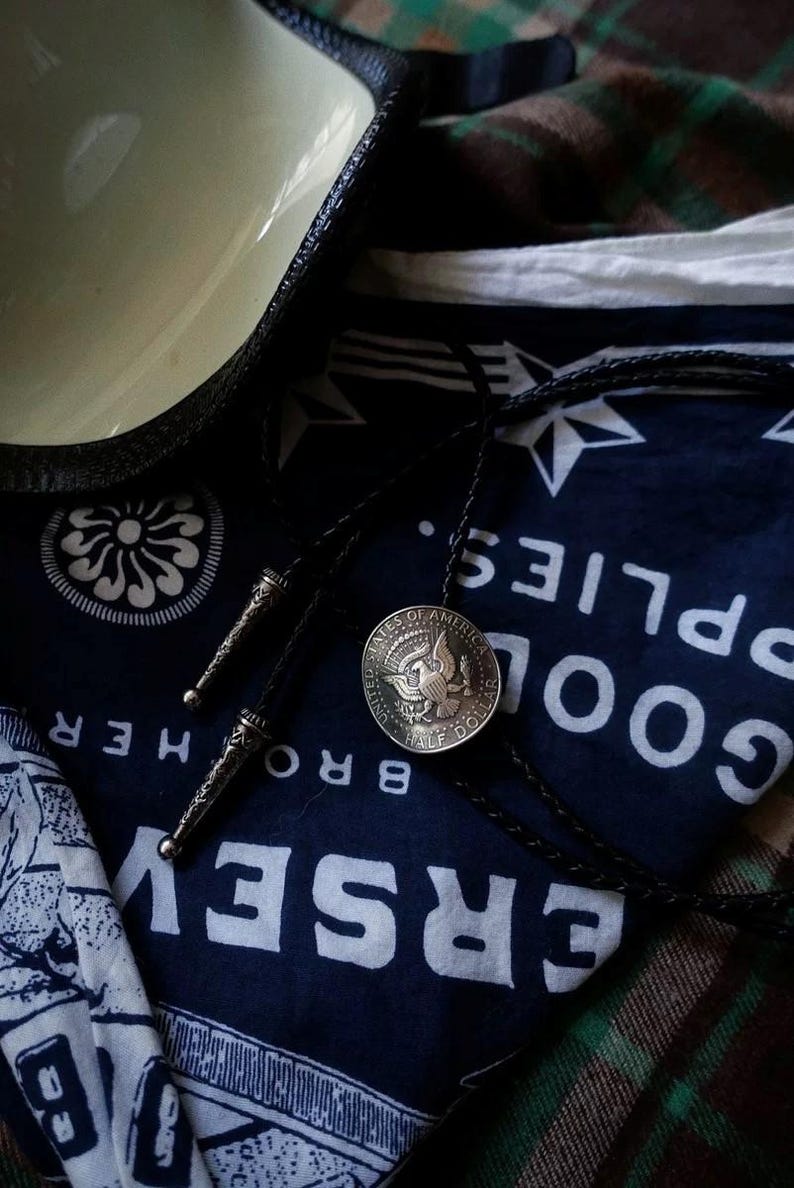 K&ouml;nnte beinhalten: Eine Bolo-Krawatte mit einer silbernen Half-Dollar-M&uuml;nze und einer schwarzen Lederschnur. Die M&uuml;nze zeigt einen Adler und die Worte "United States of America". Die Krawatte liegt auf einem marineblauen Bandana mit wei&szlig;em Text und Sternmustern.
