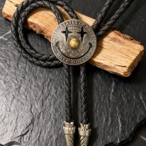 May include: A black leather bolo tie featuring a silver-toned half-dollar coin pendant with a brass center. The coin is engraved with the words "UNITED STATES OF AMERICA" and "HALF DOLLAR". The braided leather cord has decorative silver-toned tips.