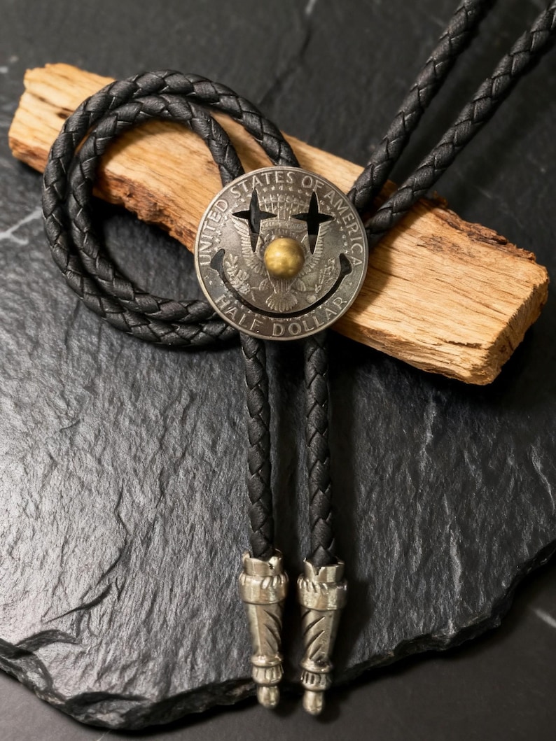 May include: A black leather bolo tie featuring a silver-toned half-dollar coin pendant with a brass center. The coin is engraved with the words "UNITED STATES OF AMERICA" and "HALF DOLLAR". The braided leather cord has decorative silver-toned tips.