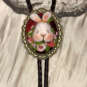 May include: A bolo tie featuring an oval antique bronze frame containing a needle-felted white rabbit head surrounded by embroidered pink and green flowers on a deep red background, attached to a braided dark brown leather cord.